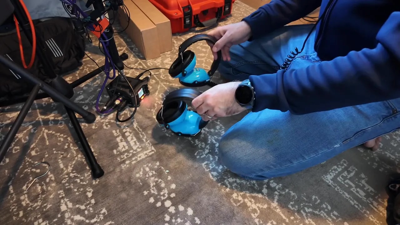 Hands holding hearing protection earmuffs above a wooden floor beside recording equipment